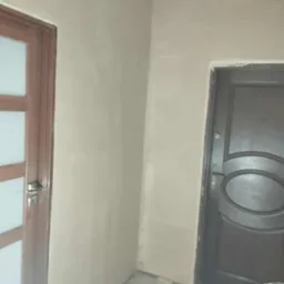 Wykończone ściany gładzią w narożniku pokoju z dwiema parami drzwi, widoczne ślady po remoncie na podłodze i drabina. Neutralne kolory ścian.