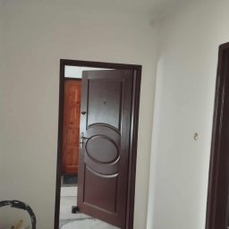 Usługi remontowo budowlane Adrian Matwiejczuk - Świeżo pomalowane, jasne ściany w korytarzu z brązowymi drzwiami i widocznym wiadrem z farbą. Minimalistyczne oświetlenie.