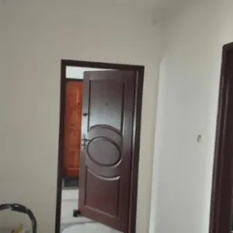 Świeżo pomalowane, jasne ściany w korytarzu z brązowymi drzwiami i widocznym wiadrem z farbą. Minimalistyczne oświetlenie.