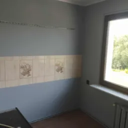 Szare ściany kuchni po malowaniu z dekoracyjnymi płytkami i listwą LED, widoczny fragment podłogi i grzejnika, okno z widokiem na zieleń.