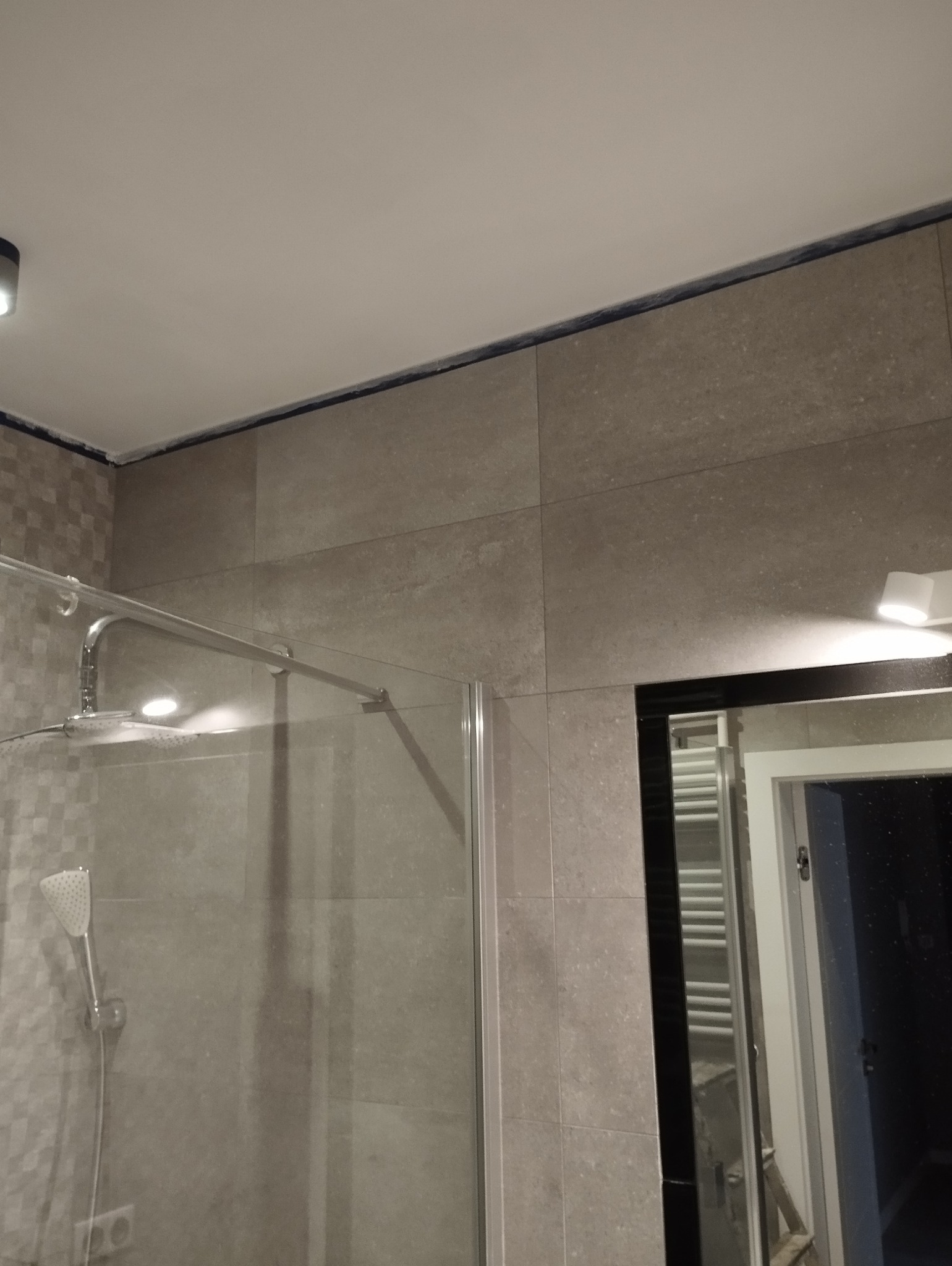 Szklana kabina prysznicowa z chromowaną armaturą i deszczownicą, obłożona beżowymi płytkami w dużej formatce, widoczna fuga silikonowa na styku ściany i sufitu, odbicie wnętrza łazienki w lustrze.