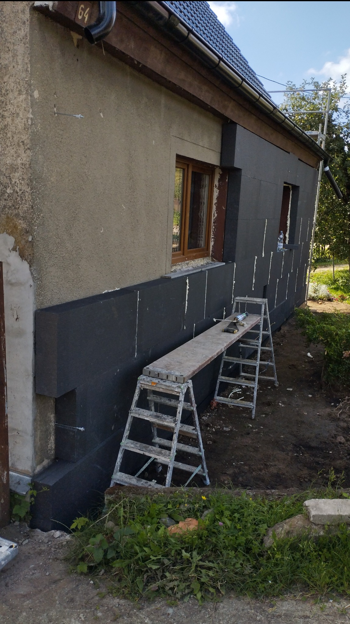 Ocieplanie elewacji budynku styropianem grafitowym, widoczne rusztowanie, częściowo otynkowana ściana i numer domu '64' nad oknem.