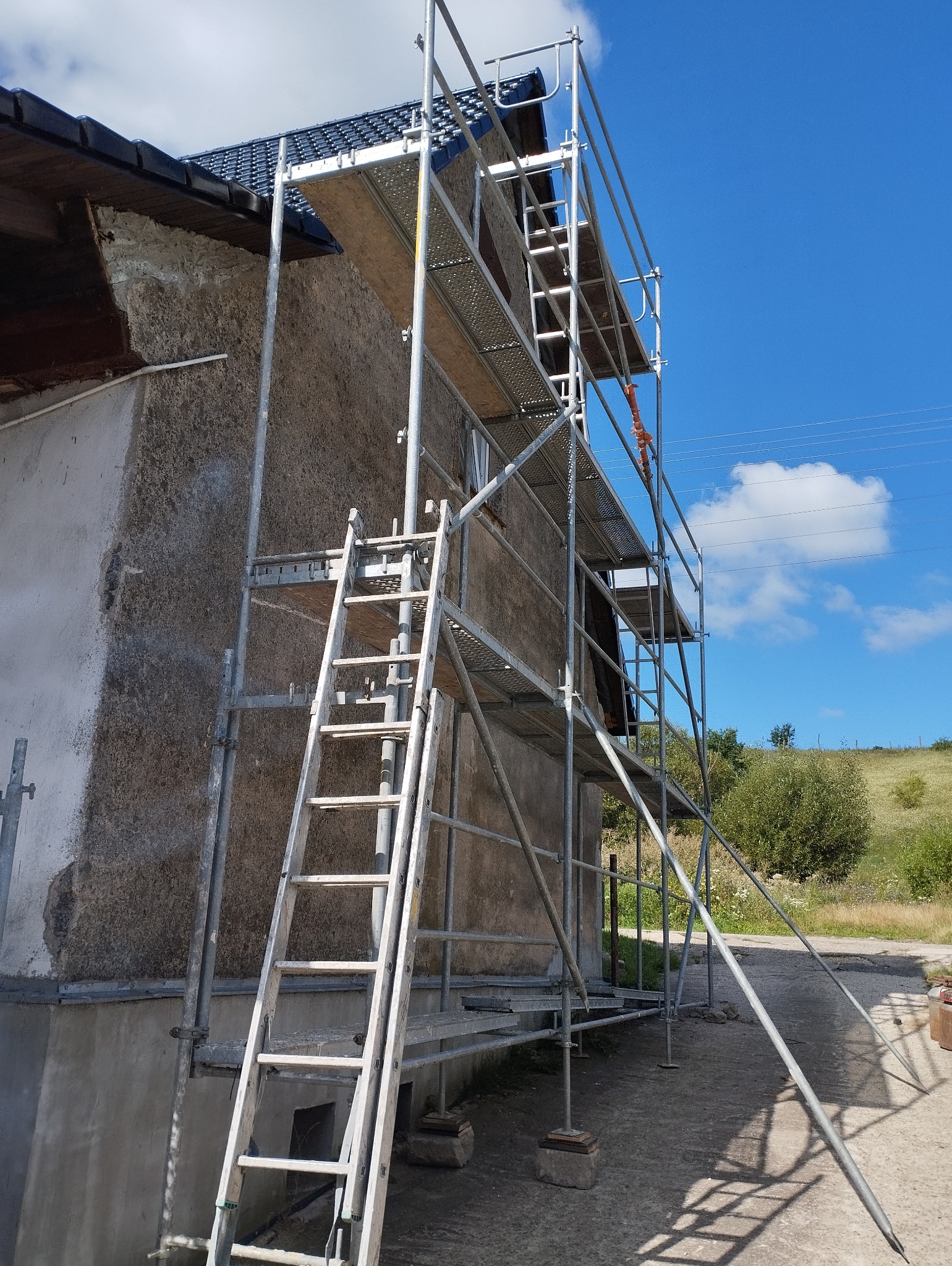 Metalowe rusztowanie przy szarej ścianie budynku z widocznymi śladami napraw, na tle błękitnego nieba z chmurami i zielenią wzgórz.