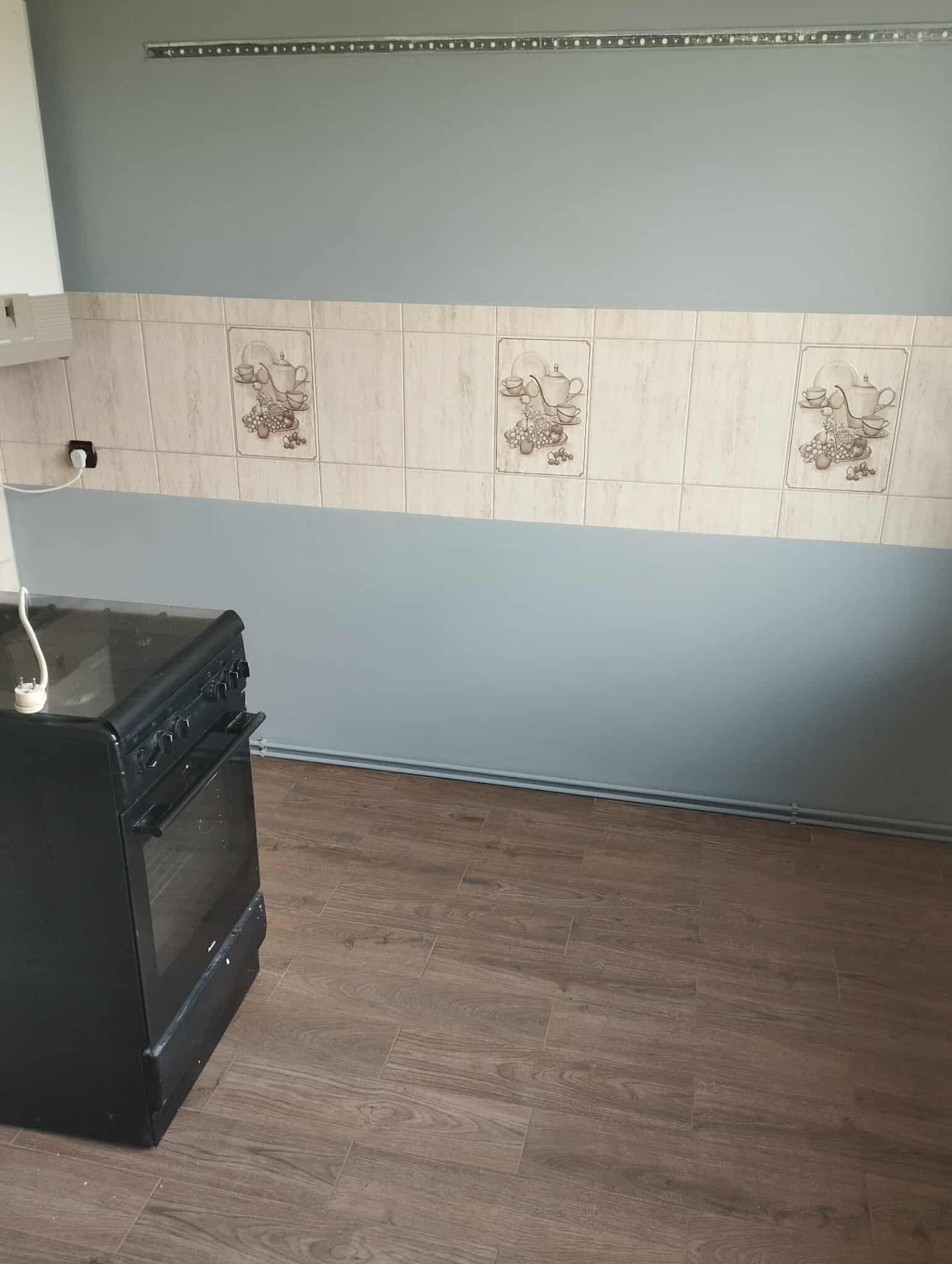 Fragment kuchni po remoncie: świeżo pomalowana ściana w odcieniach szarości, ułożone płytki z motywem filiżanek i owoców, podłoga z paneli imitujących drewno.