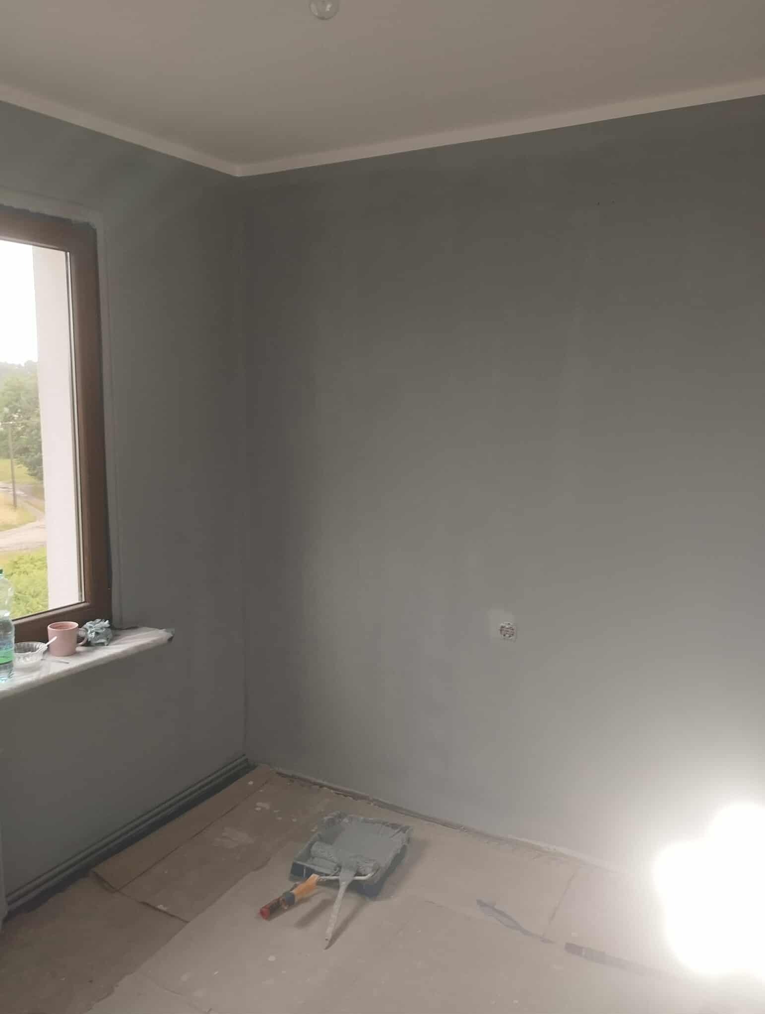 Pomieszczenie po malowaniu ścian na szaro, widoczny wałek i kuweta z farbą. Okno z widokiem na zewnątrz. Listwa pod sufitem. Podłoga zabezpieczona kartonem.