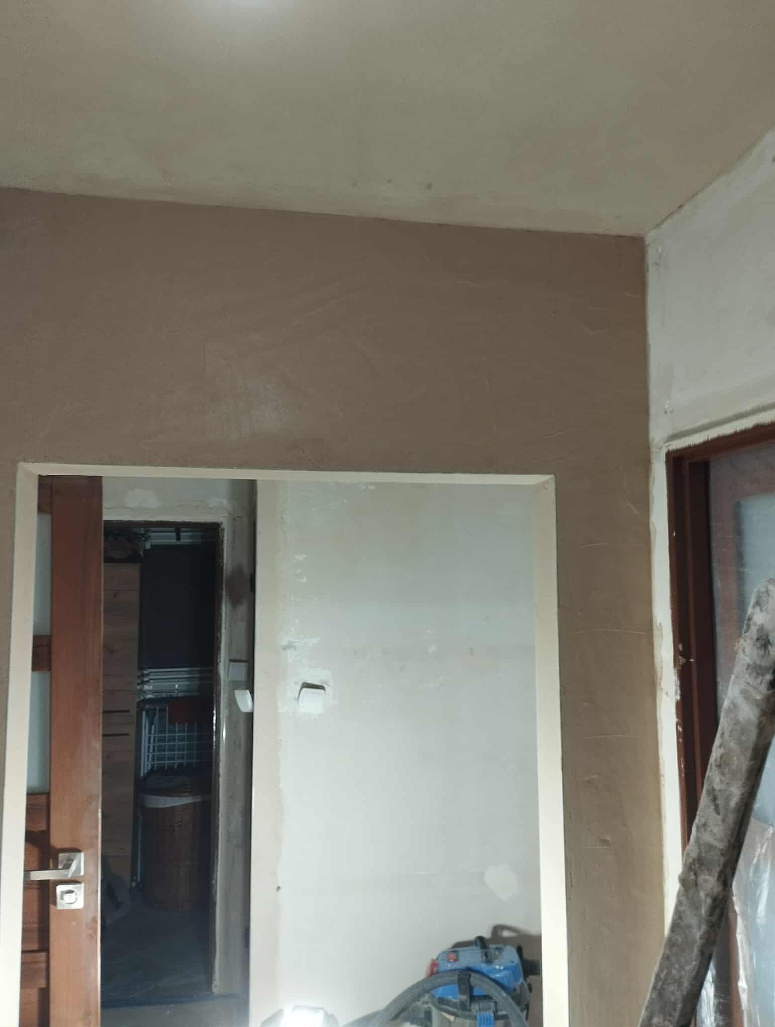 Ściana po nałożeniu gładzi w odcieniach beżu, widoczne ślady szpachlowania, w tle drzwi do innego pomieszczenia i drabina oparta o ścianę.