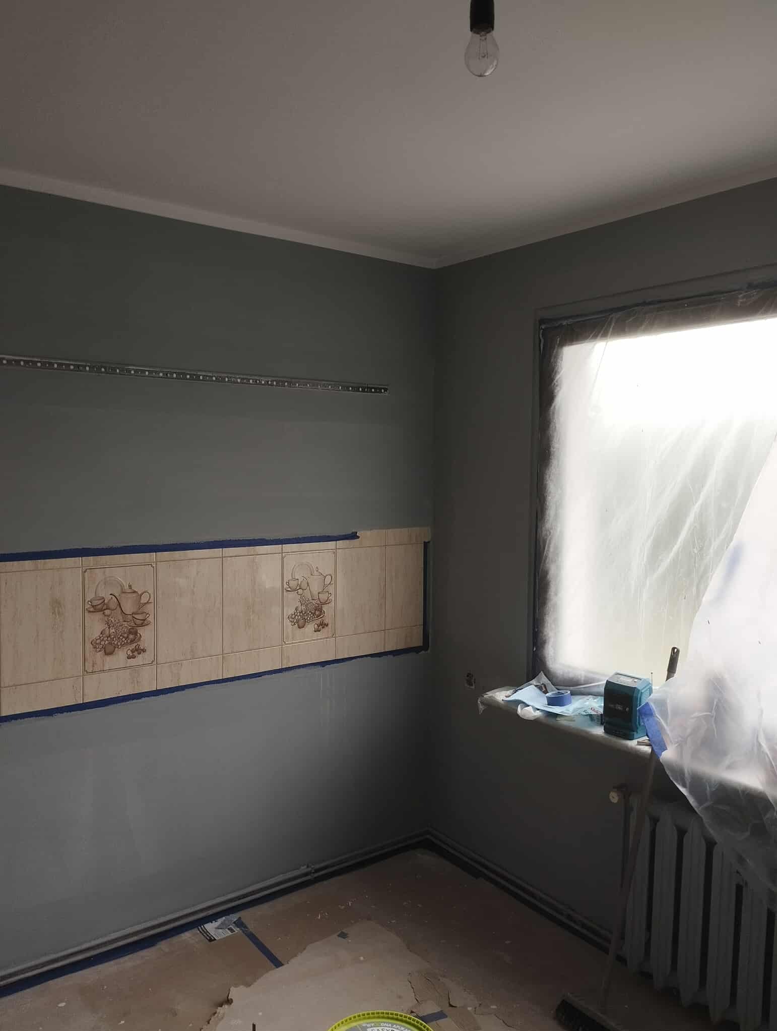 Pokój w trakcie remontu: szare ściany, częściowo usunięte płytki z motywem filiżanek, okno zakryte folią, widoczny grzejnik i surowe podłoże.