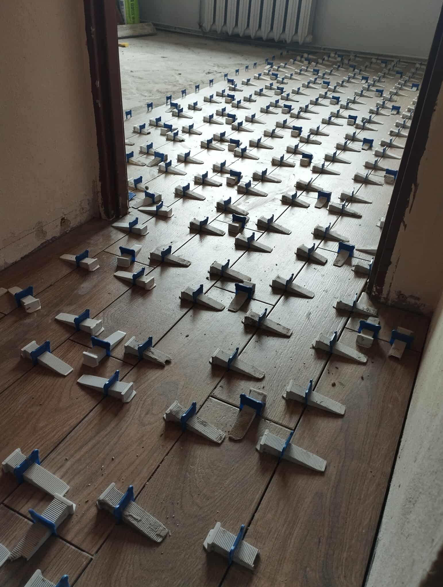 Układanie płytek podłogowych z użyciem klipsów poziomujących. Widoczne brązowe płytki imitujące drewno i biało-niebieskie klipsy. Próg drzwiowy w tle.