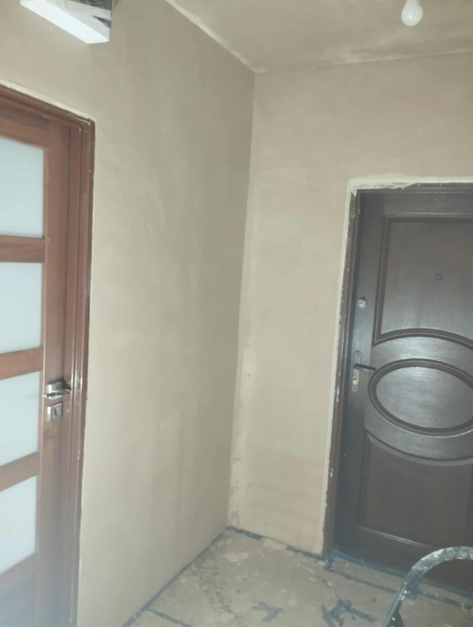 Wykończone ściany gładzią w narożniku pokoju z dwiema parami drzwi, widoczne ślady po remoncie na podłodze i drabina. Neutralne kolory ścian.