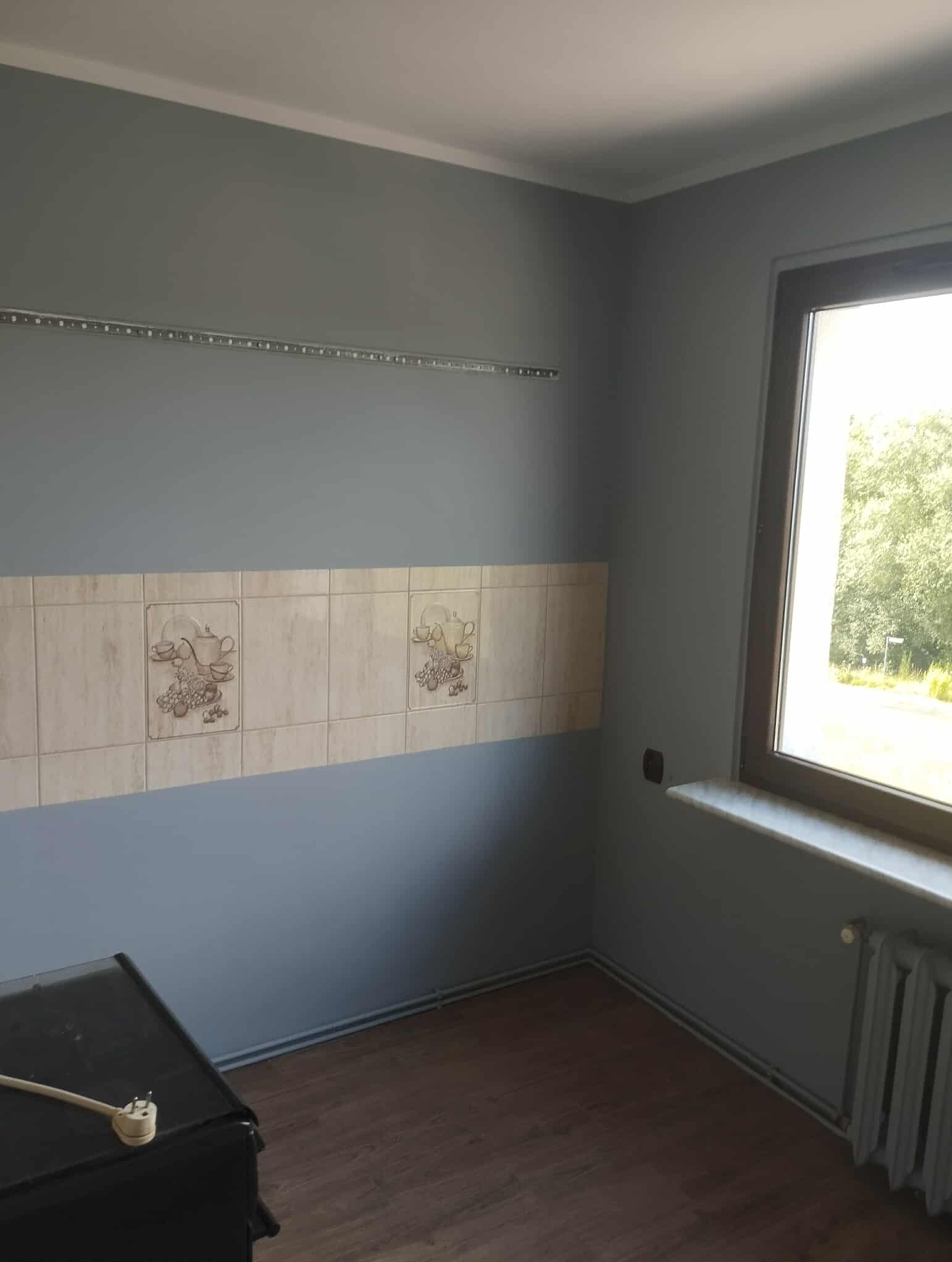 Szare ściany kuchni po malowaniu z dekoracyjnymi płytkami i listwą LED, widoczny fragment podłogi i grzejnika, okno z widokiem na zieleń.