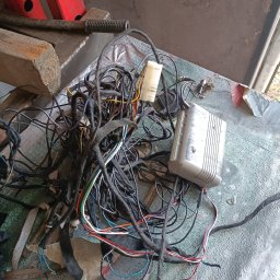 QuickService Mateusz Góra - Skomplikowana wiązka przewodów samochodowych z kostką i sterownikiem leżąca na brudnym stole warsztatowym, w tle widoczny podnośnik warsztatowy.