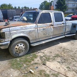 QuickService Mateusz Góra - Pickup Ford F-350 w trakcie renowacji blacharskiej, widoczne ślady korozji, szlifowania i brakujące elementy karoserii na tle podwórka z innymi pojazdami.