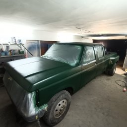 QuickService Mateusz Góra - Pick-up w trakcie lakierowania na ciemnozielony kolor, zabezpieczony folią i taśmą malarską, widoczne półki z farbami w tle warsztatu.