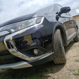 QuickService Mateusz Góra - Częściowy widok czarnego SUV-a marki Mitsubishi Outlander z brudnymi oponami na szutrowym podłożu, z odbiciem fotografa w lakierze.