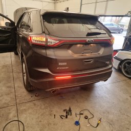 QuickService Mateusz Góra - Brązowy Ford Edge z otwartą klapą bagażnika i włączonymi światłami pozycyjnymi, widoczne narzędzia i przewody na podłodze warsztatu, sugerujące prace elektryczne przy pojeździe.