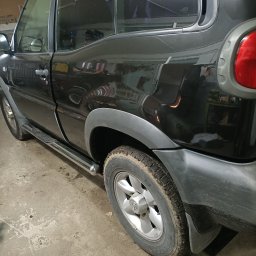 QuickService Mateusz Góra - Częściowy widok boku czarnego samochodu terenowego z aluminiowymi felgami, lekko zabrudzonymi oponami i chromowanym progiem, zaparkowanego w garażu z betonową podłogą.