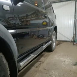 Czarny samochód terenowy z chromowanymi progami w warsztacie, widoczne opony terenowe i fragment bramy garażowej.