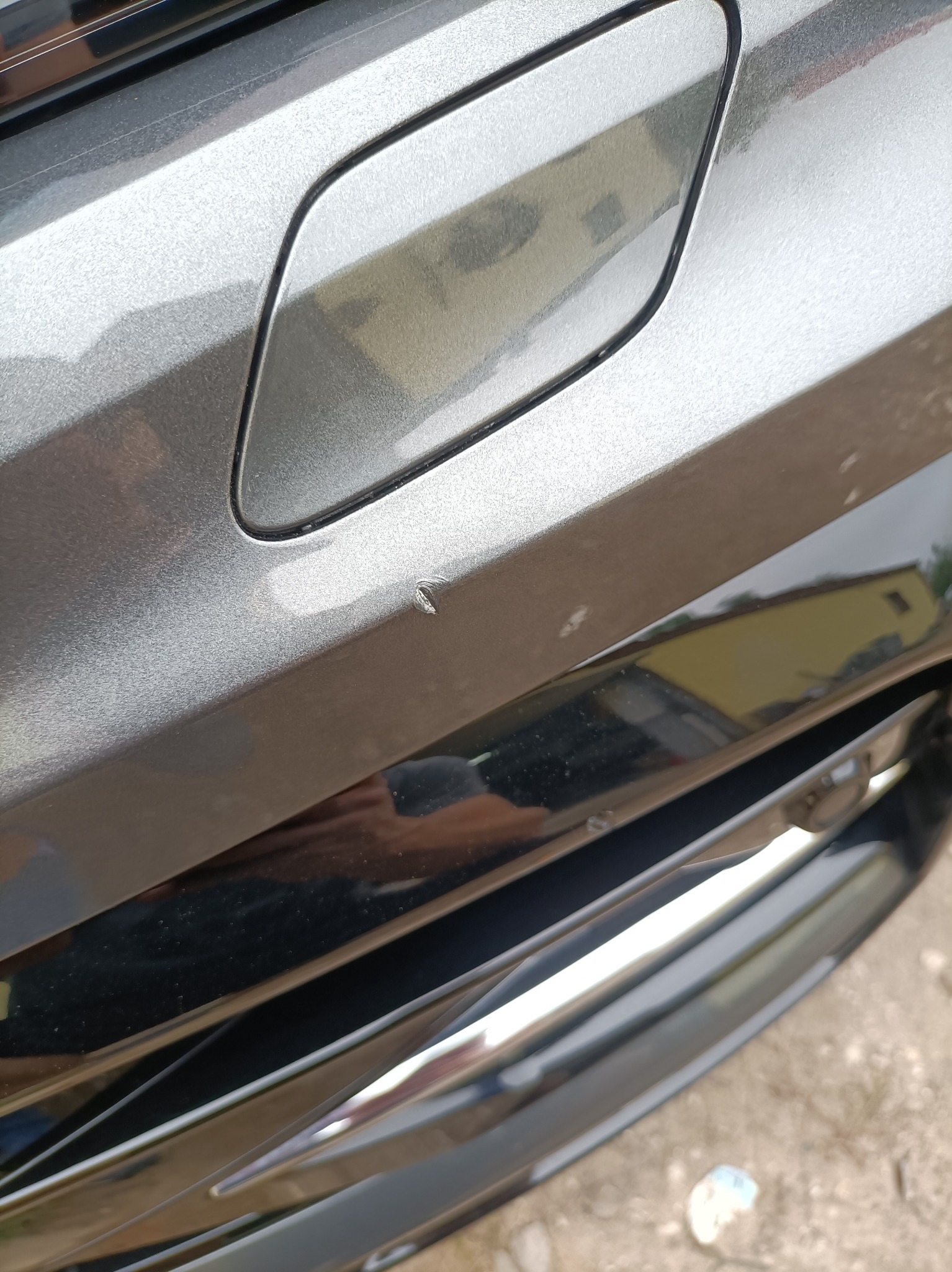 Niewielkie uszkodzenie lakieru na srebrnym błotniku samochodu, widoczny odprysk i zarysowania. W tle odbicie budynku.