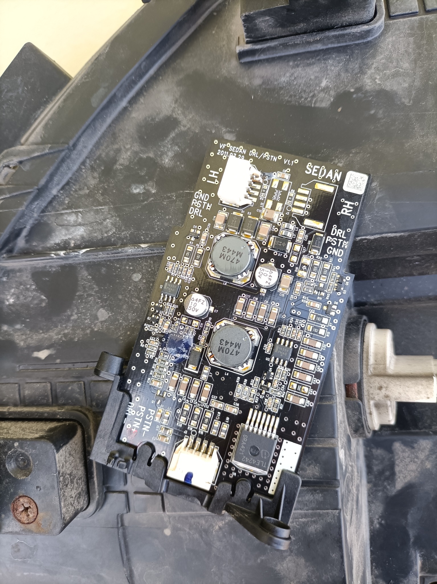 Uszkodzona płyta sterująca LED DRL w reflektorze samochodowym typu Sedan, widoczne kondensatory, rezystory i układy scalone, zabrudzona obudowa reflektora.