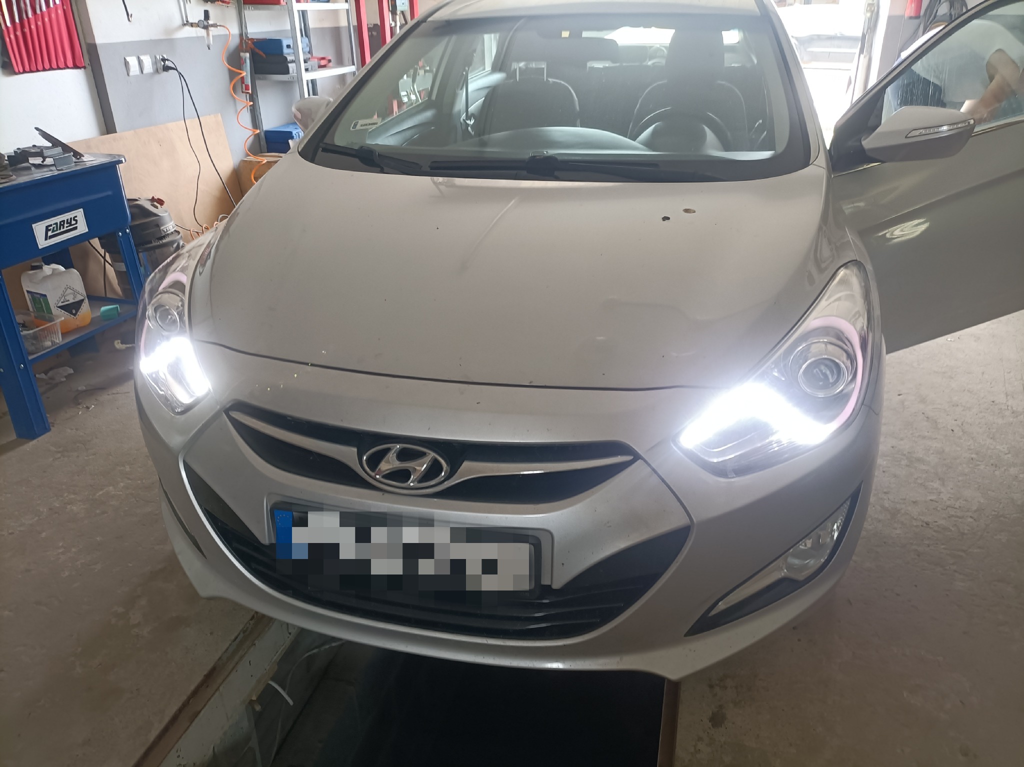 Przód srebrnego samochodu Hyundai z włączonymi światłami LED na stanowisku warsztatowym z otwartymi drzwiami kierowcy.
