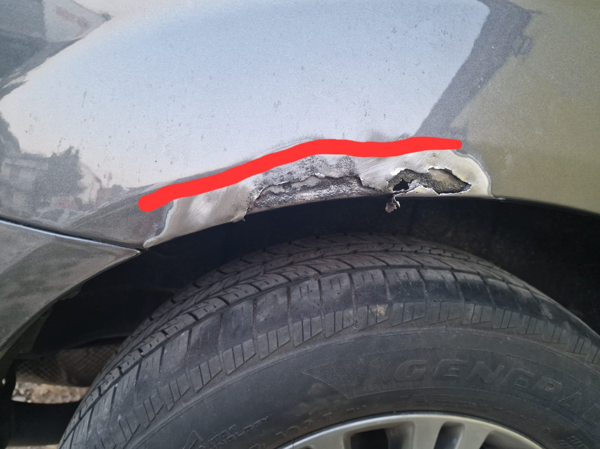 Uszkodzony, skorodowany błotnik samochodu w kolorze szarym, widoczna rdza i ubytki blachy nad oponą.