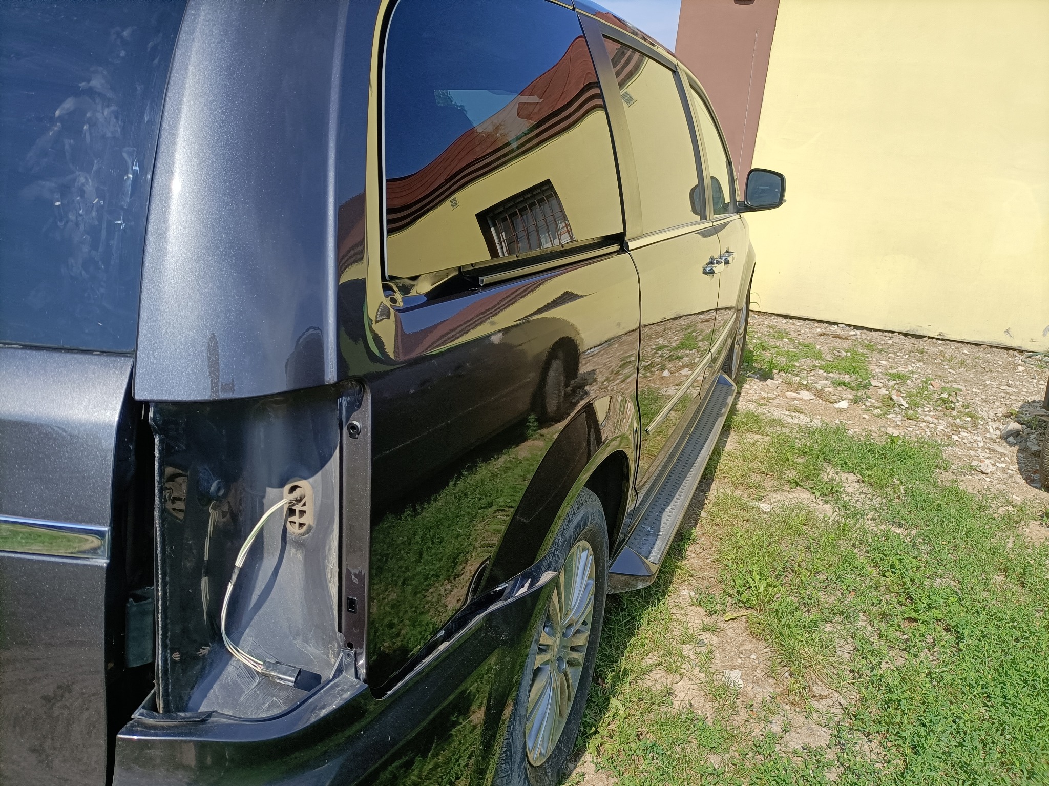 Szary samochód z uszkodzonym tyłem, brakuje lampy i widać przewody. W tle budynek z żółtą ścianą i odbicie w szybie.