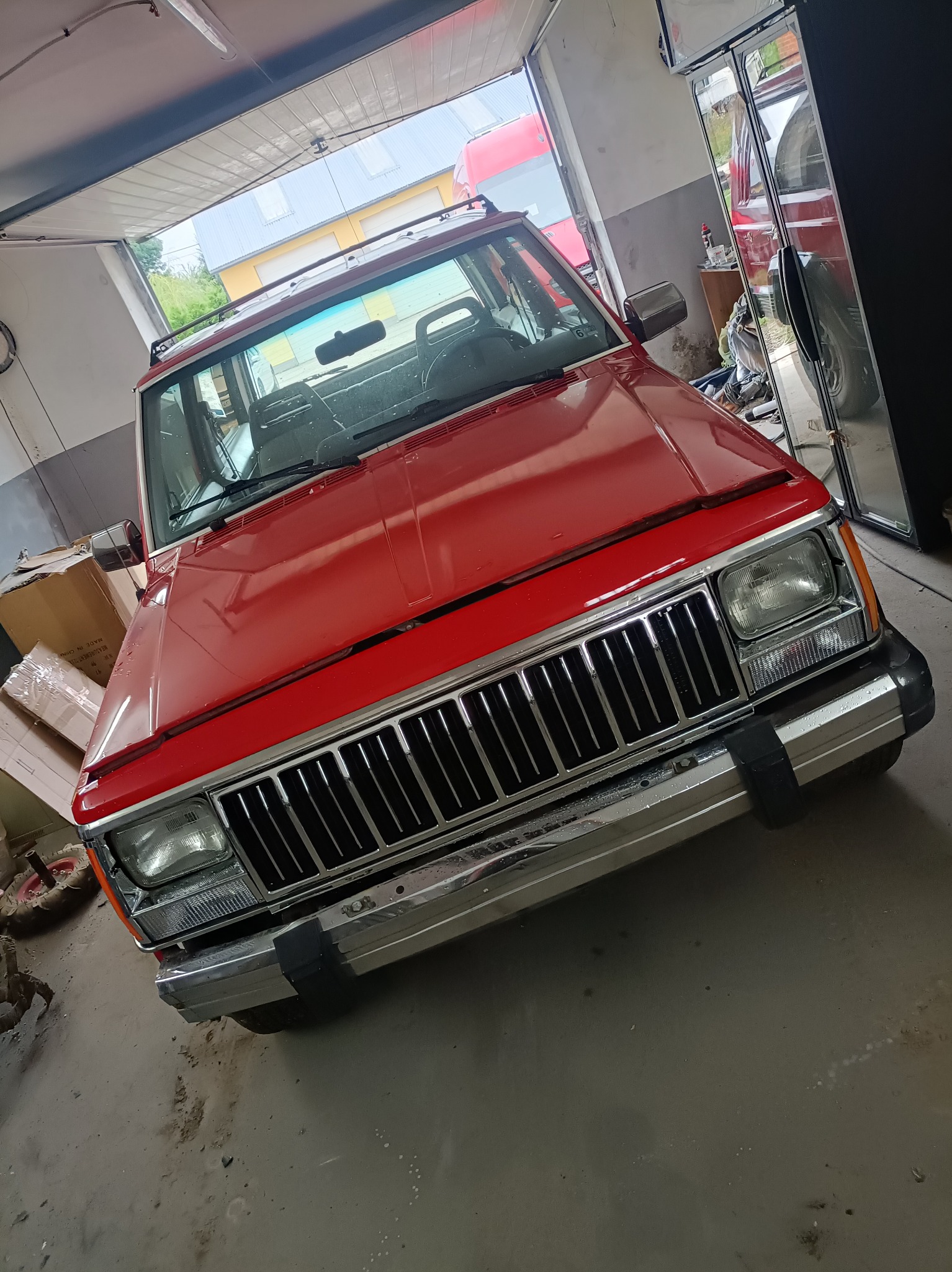 Czerwony samochód Jeep Cherokee w warsztacie, widok z przodu, na masce widoczne krople wody, w tle otwarte drzwi garażowe i inne pojazdy.