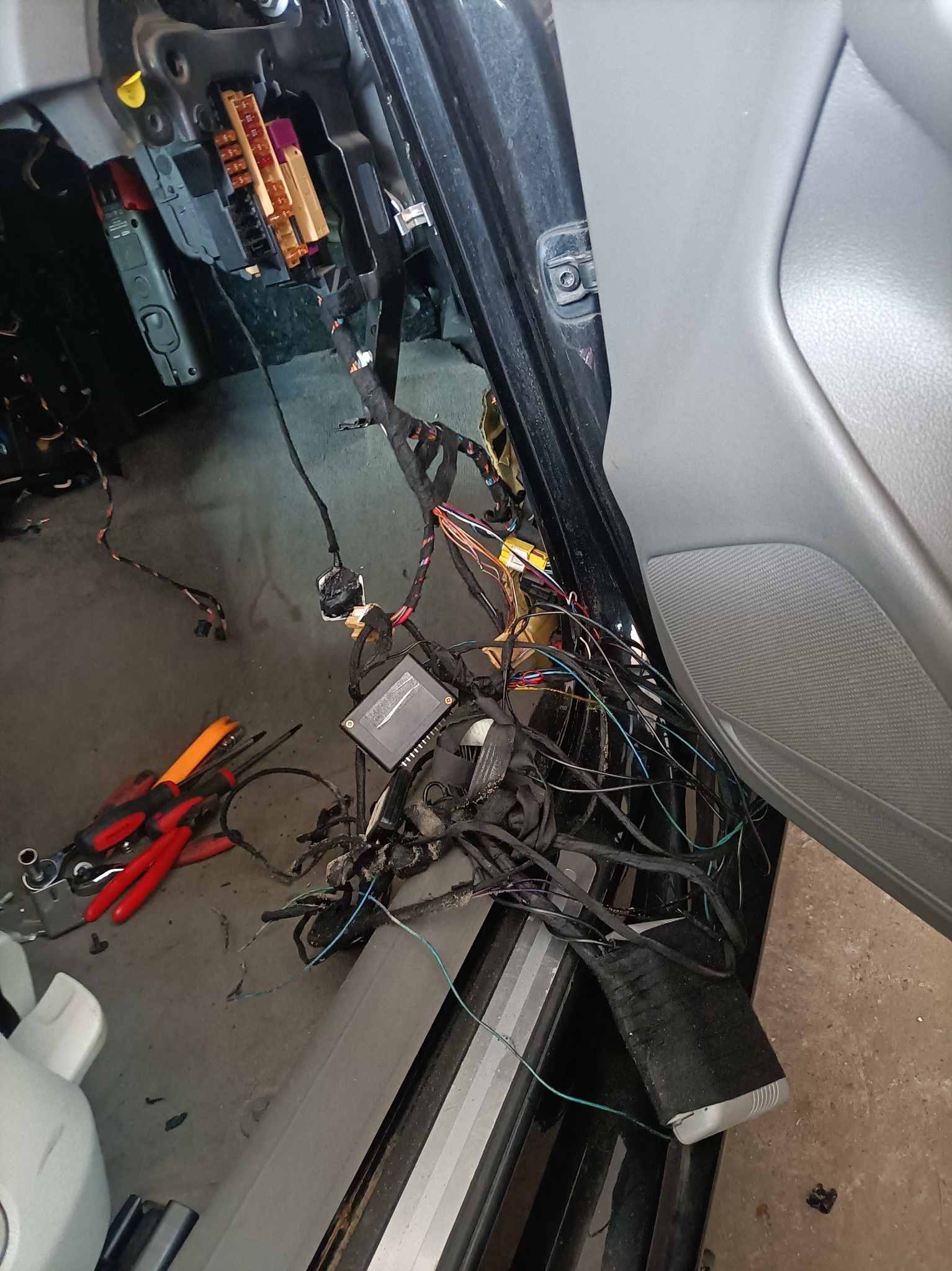 Otwarta skrzynka bezpieczników samochodowych z widocznymi przewodami, narzędzia i elementy wnętrza pojazdu podczas naprawy.