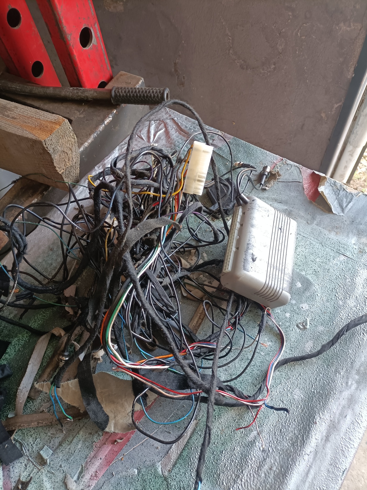 Skomplikowana wiązka przewodów samochodowych z kostką i sterownikiem leżąca na brudnym stole warsztatowym, w tle widoczny podnośnik warsztatowy.