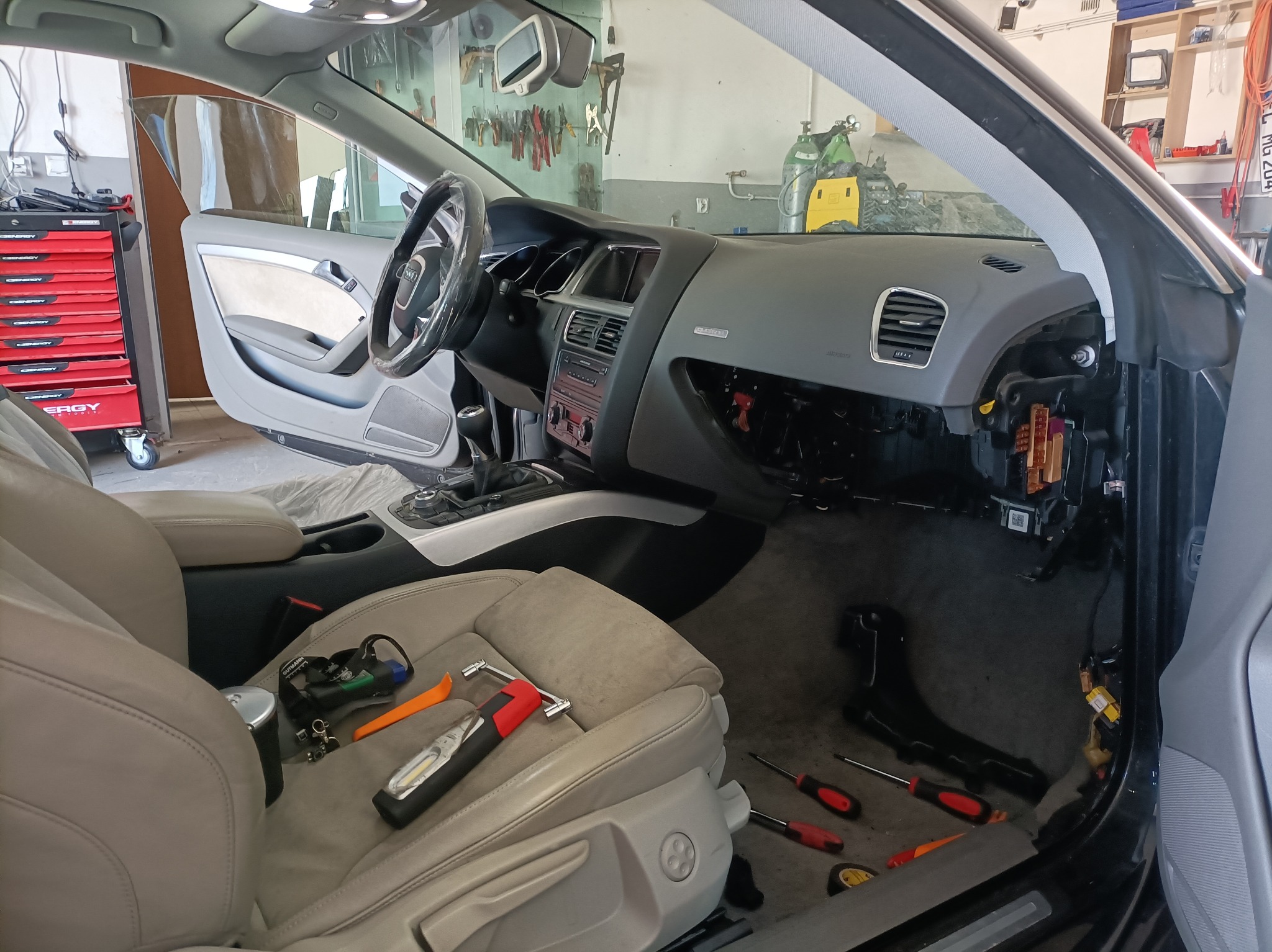 Wnętrze samochodu Audi A5 w trakcie demontażu deski rozdzielczej, widoczne narzędzia na siedzeniu i podłodze, odsłonięte elementy instalacji elektrycznej.