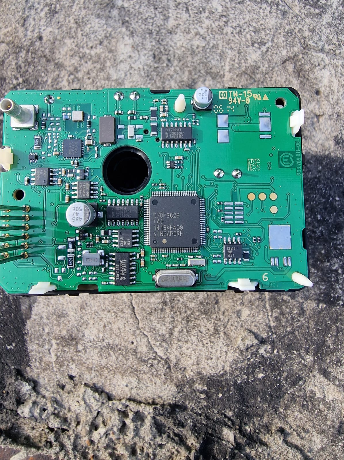 Zielona płytka drukowana z widocznymi elementami elektronicznymi, układami scalonymi i ścieżkami, umieszczona na szarej, betonowej powierzchni.