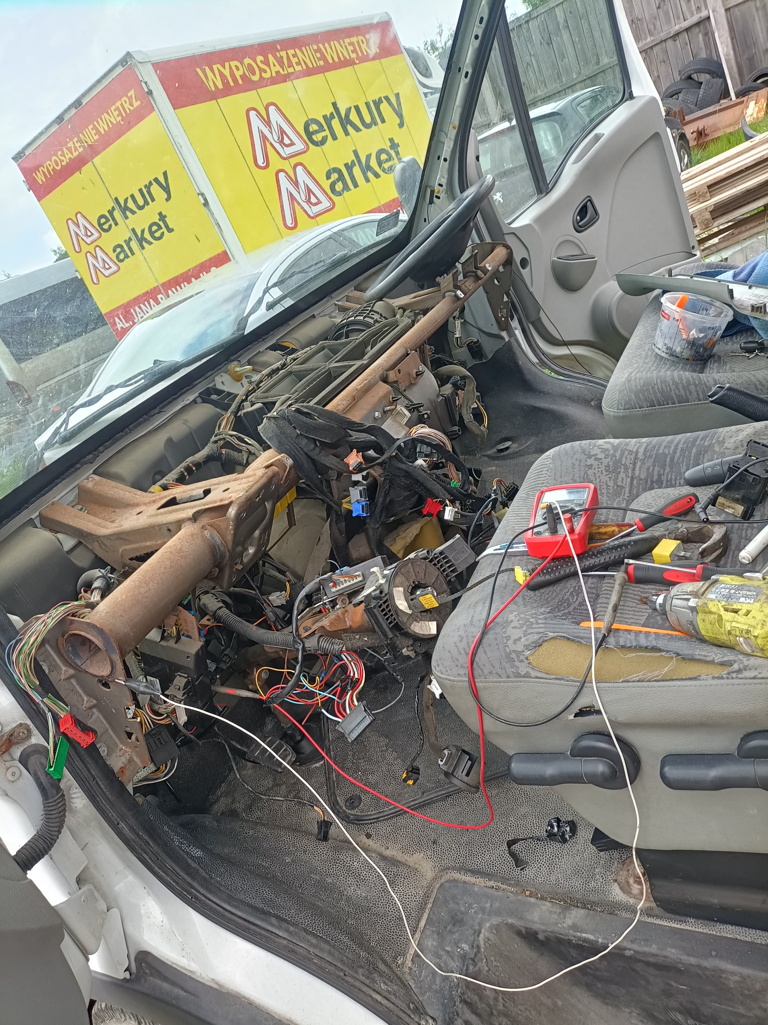 Wnętrze kabiny dostawczego samochodu z widoczną zdemontowaną deską rozdzielczą, wiązkami kabli i narzędziami na siedzeniu, w tle żółty samochód dostawczy Merkury Market.