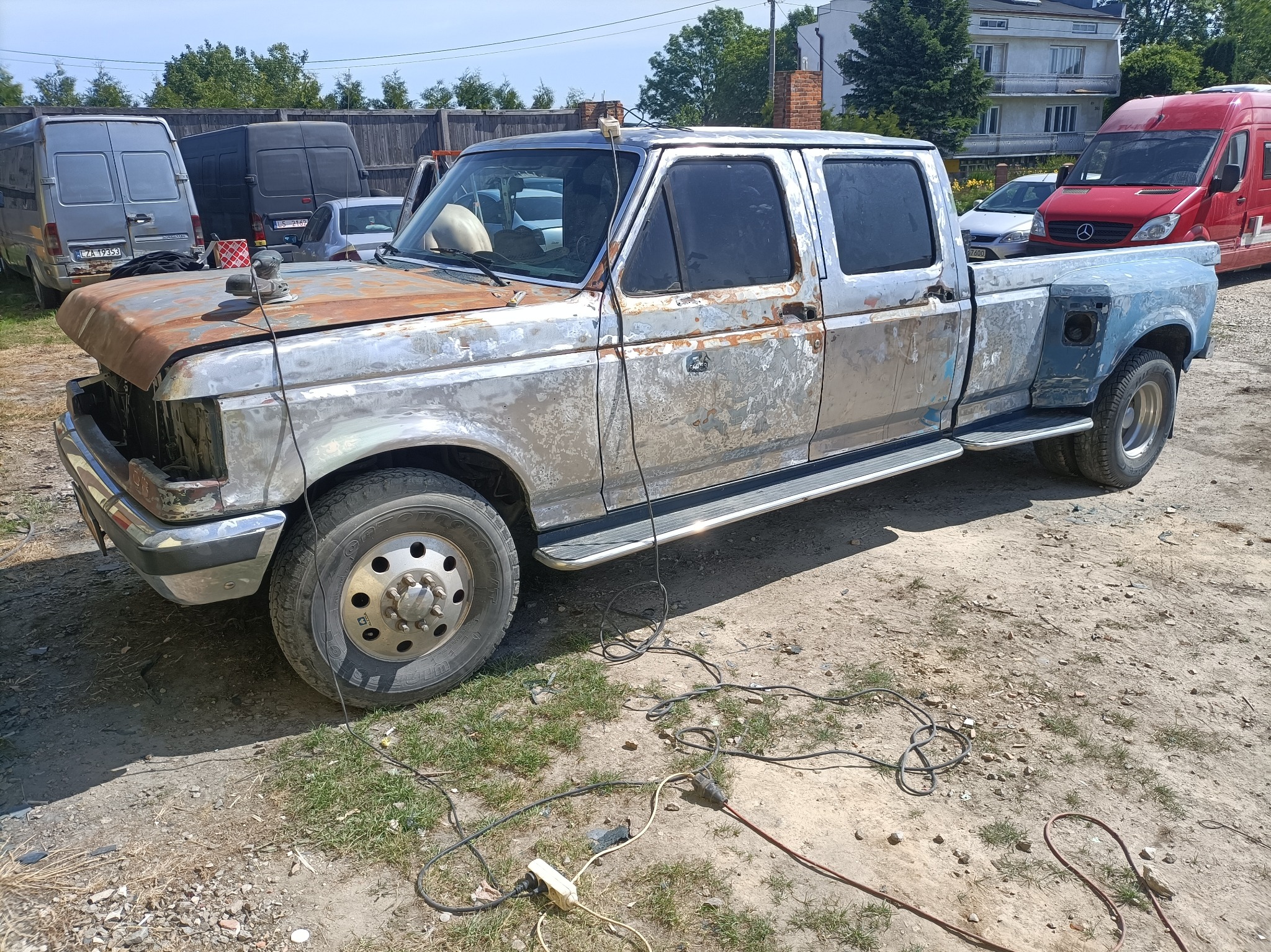 Pickup Ford F-350 w trakcie renowacji blacharskiej, widoczne ślady korozji, szlifowania i brakujące elementy karoserii na tle podwórka z innymi pojazdami.