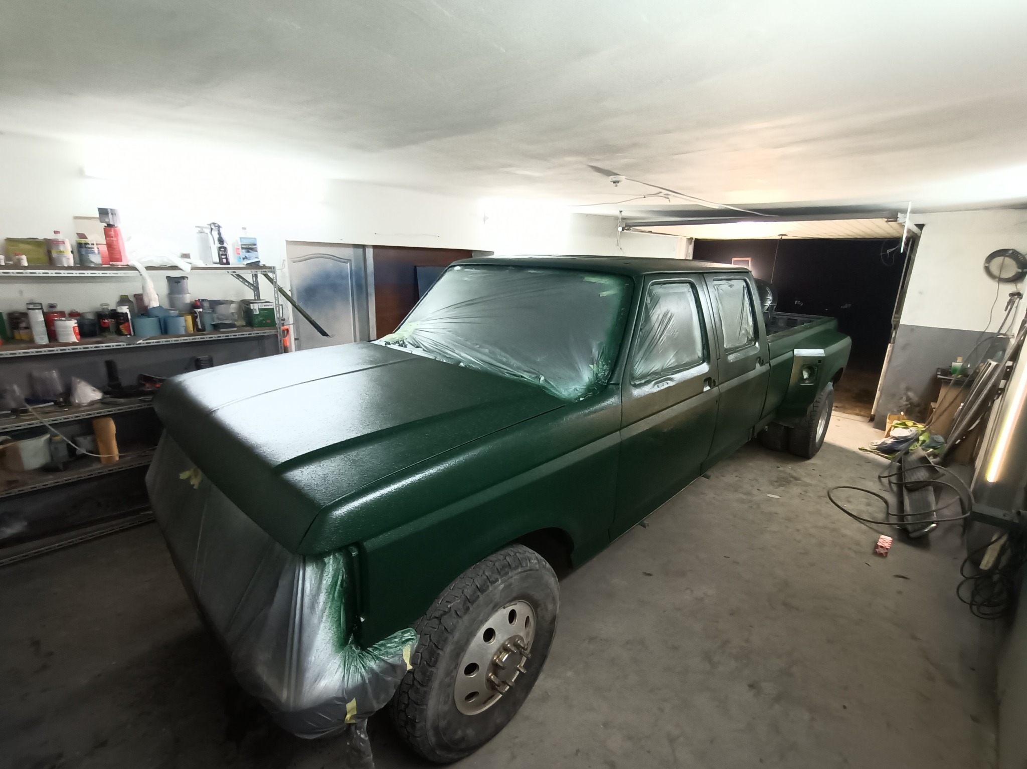 Pick-up w trakcie lakierowania na ciemnozielony kolor, zabezpieczony folią i taśmą malarską, widoczne półki z farbami w tle warsztatu.