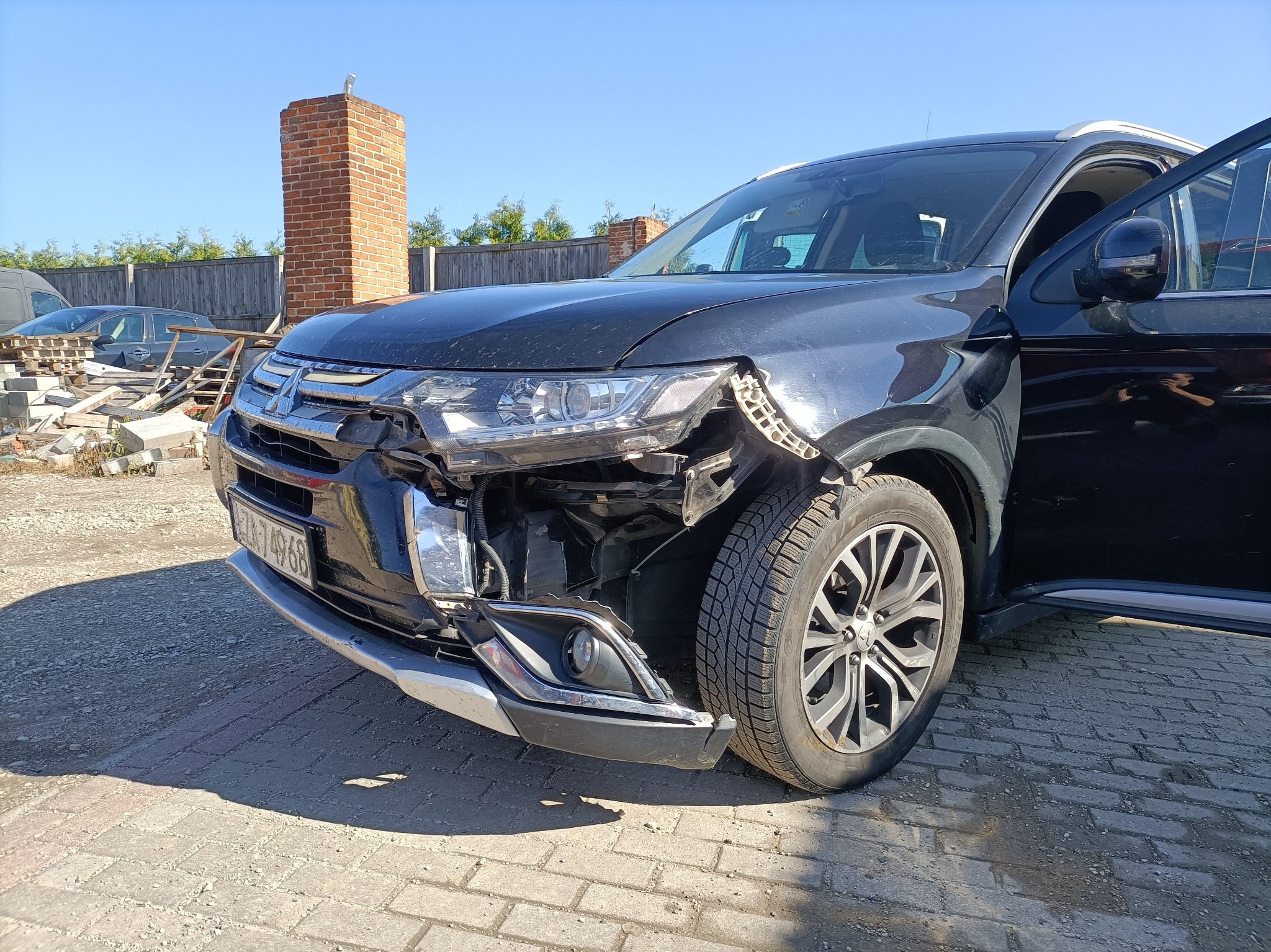Uszkodzony przód czarnego SUV-a Mitsubishi Outlander z otwartymi drzwiami kierowcy, widoczne zniszczenia reflektora i zderzaka na tle ceglanego komina i innych pojazdów.