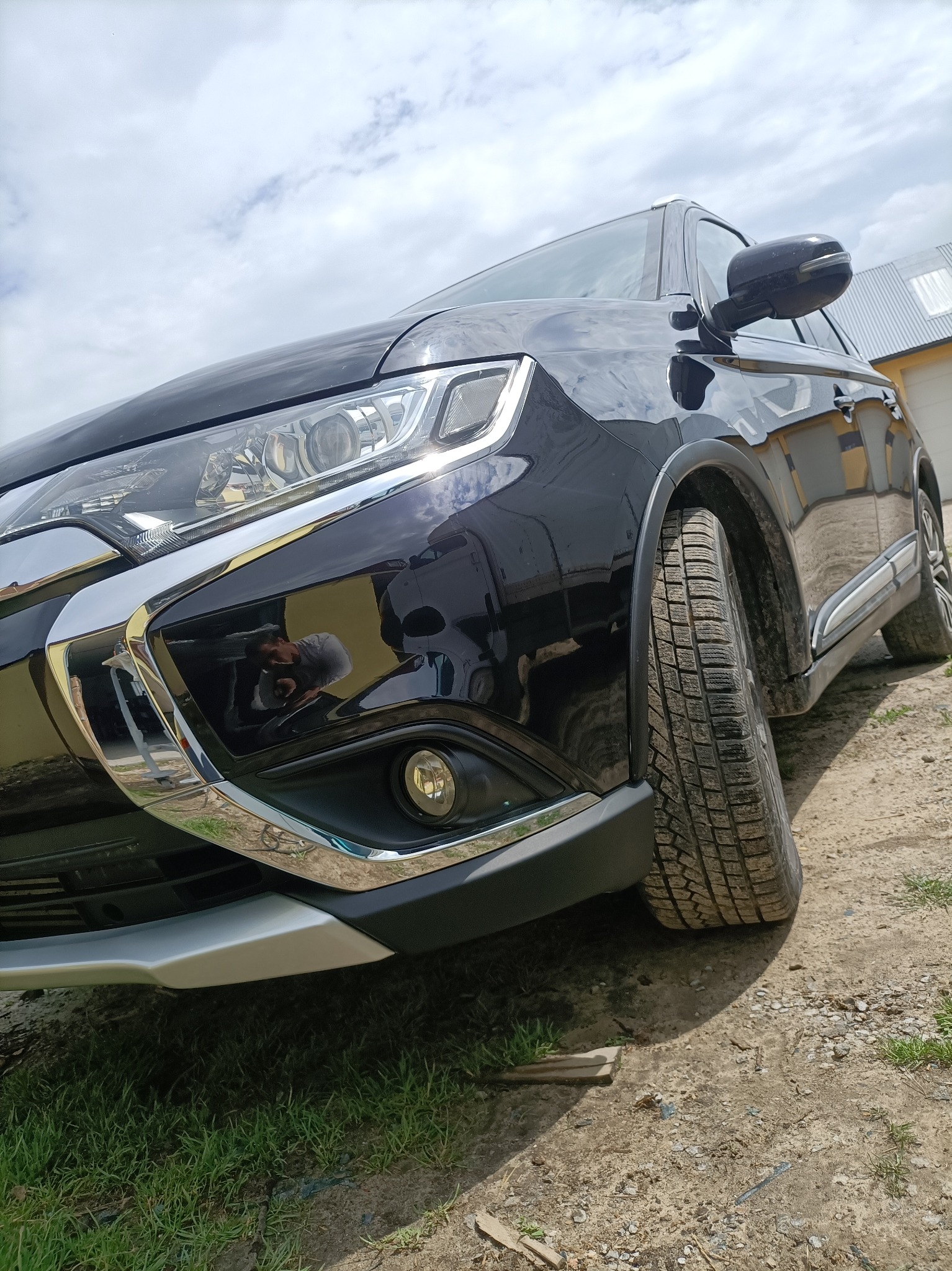 Częściowy widok czarnego SUV-a marki Mitsubishi Outlander z brudnymi oponami na szutrowym podłożu, z odbiciem fotografa w lakierze.