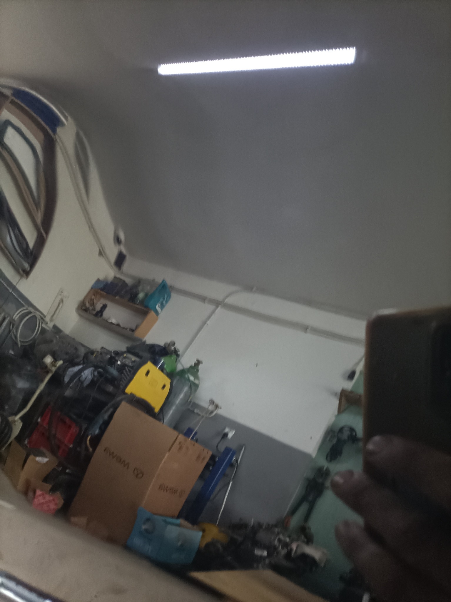 Odbicie w karoserii samochodu, ukazujące warsztat z narzędziami, częściami zamiennymi i kartonami, oświetlony jarzeniówką.