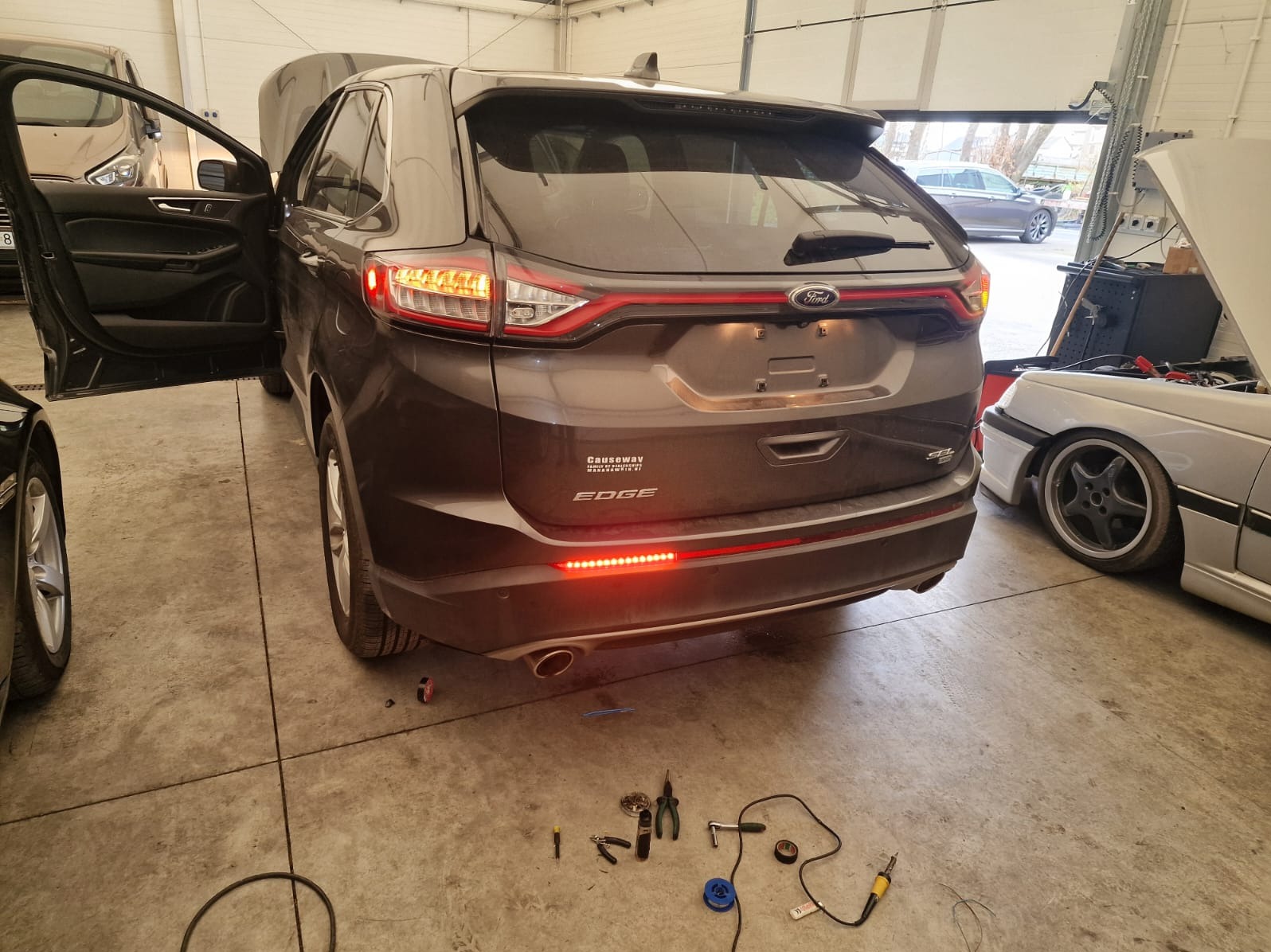 Brązowy Ford Edge z otwartą klapą bagażnika i włączonymi światłami pozycyjnymi, widoczne narzędzia i przewody na podłodze warsztatu, sugerujące prace elektryczne przy pojeździe.