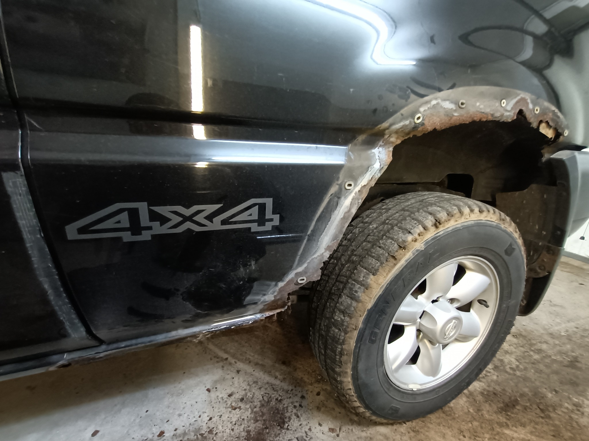 Fragment czarnego samochodu 4x4 z widocznymi uszkodzeniami i korozją nadkola, przygotowywanego do naprawy blacharskiej. Widoczne otwory po nitach lub spawach punktowych.
