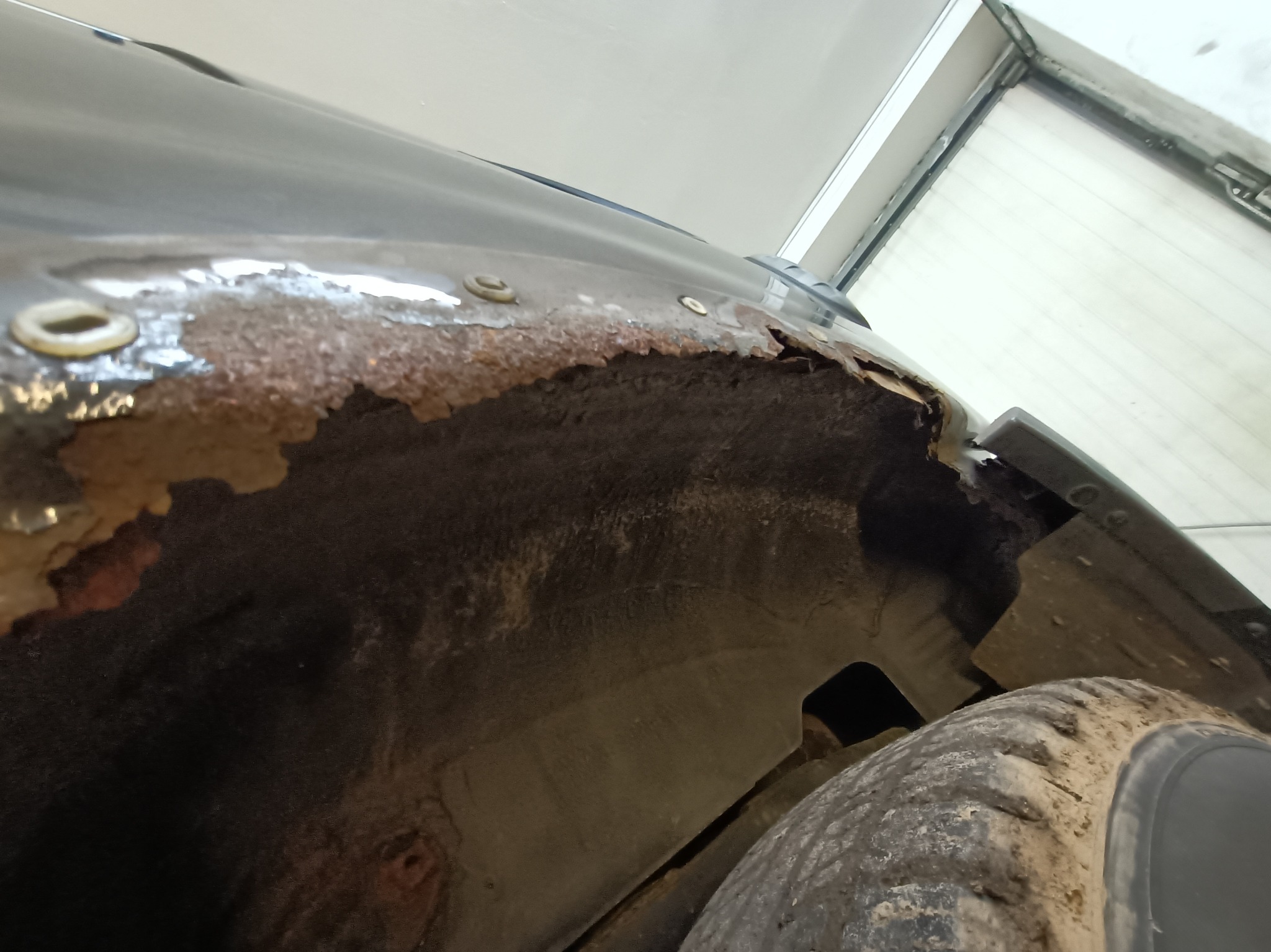 Zardzewiałe nadkole samochodu z widocznymi śladami korozji i mocowaniami, ujęcie od dołu, fragment opony w kadrze.