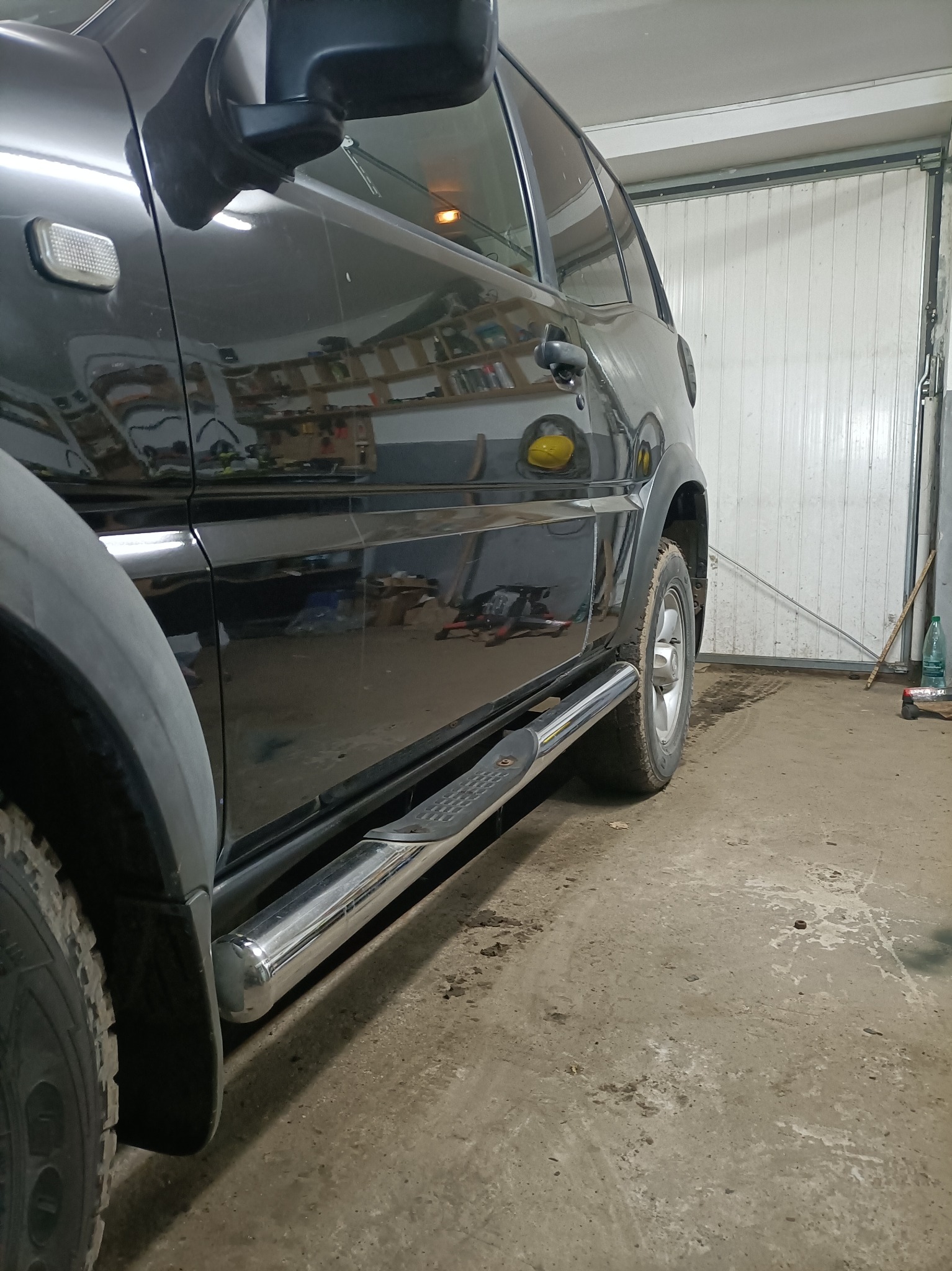 Czarny samochód terenowy z chromowanymi progami w warsztacie, widoczne opony terenowe i fragment bramy garażowej.