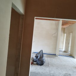 Tynki maszynowe Zdanukiewicz - Wnętrze w trakcie prac wykończeniowych, widoczne ściany po nałożeniu tynku, podłoga betonowa z niebieskimi rurami, śmieci w czarnym worku, surowy stan z widoczną konstrukcją drewnianą stropu.