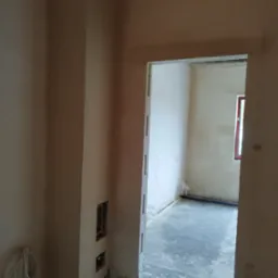 Wnętrze w trakcie prac wykończeniowych, ściany pokryte świeżym tynkiem, widoczne przewody elektryczne i otwory instalacyjne w ścianie, surowy beton na podłodze, okno w czerwonej ramie w tle.