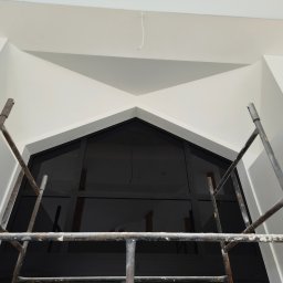 Tynki maszynowe Zdanukiewicz - Białe ściany po malowaniu, widoczne rusztowanie malarskie i okno w kształcie trójkąta. Minimalistyczne wnętrze po remoncie, z kablem zwisającym z sufitu.