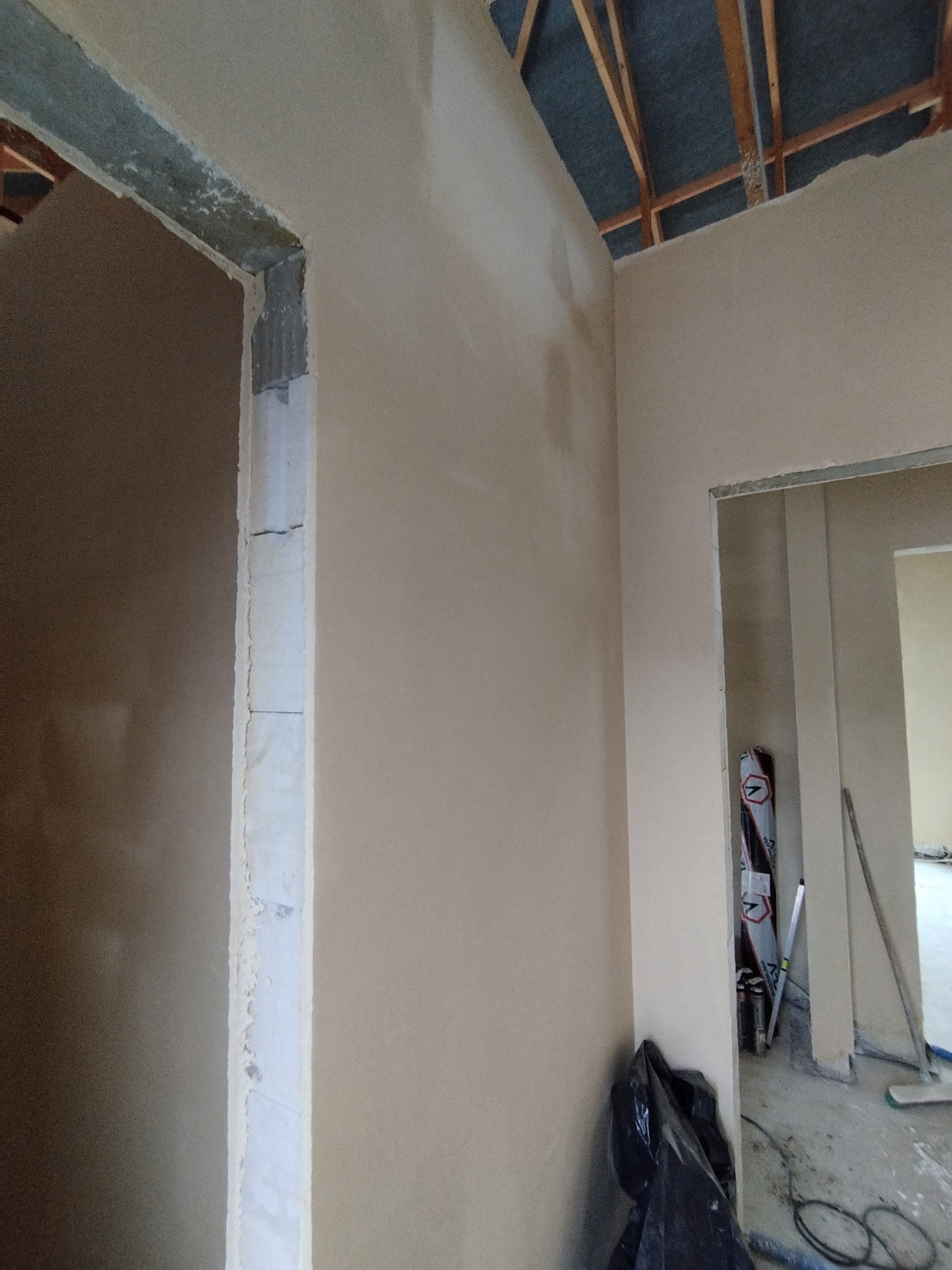 Wnętrze w trakcie prac wykończeniowych: ściany pokryte świeżo nałożoną, jasną warstwą, widoczne surowe drewniane elementy konstrukcyjne pod sufitem, w tle narzędzia i materiały budowlane.