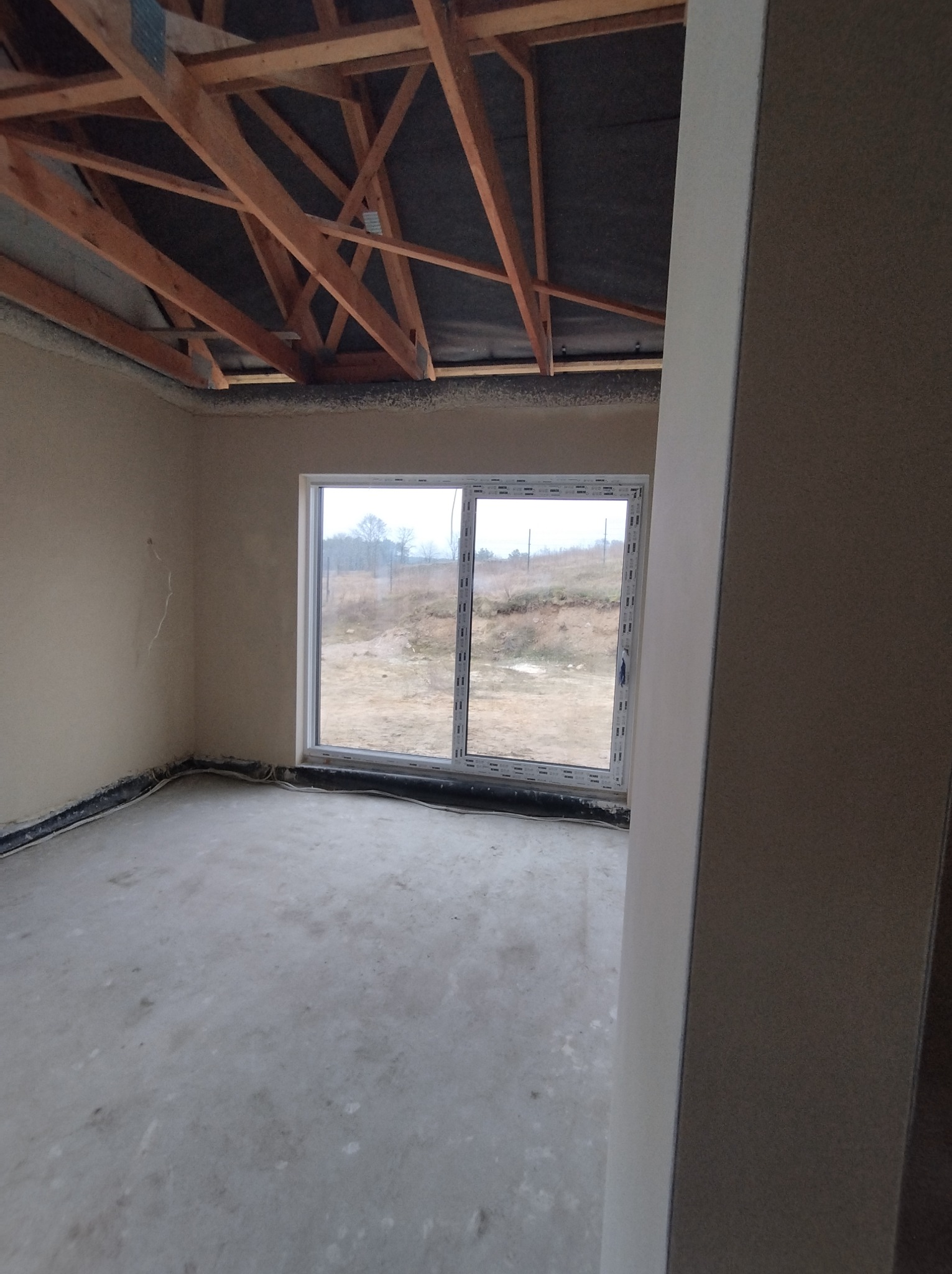Surowe wnętrze domu w trakcie budowy, ściany wstępnie pokryte tynkiem, widoczna konstrukcja dachu i okno z widokiem na krajobraz.