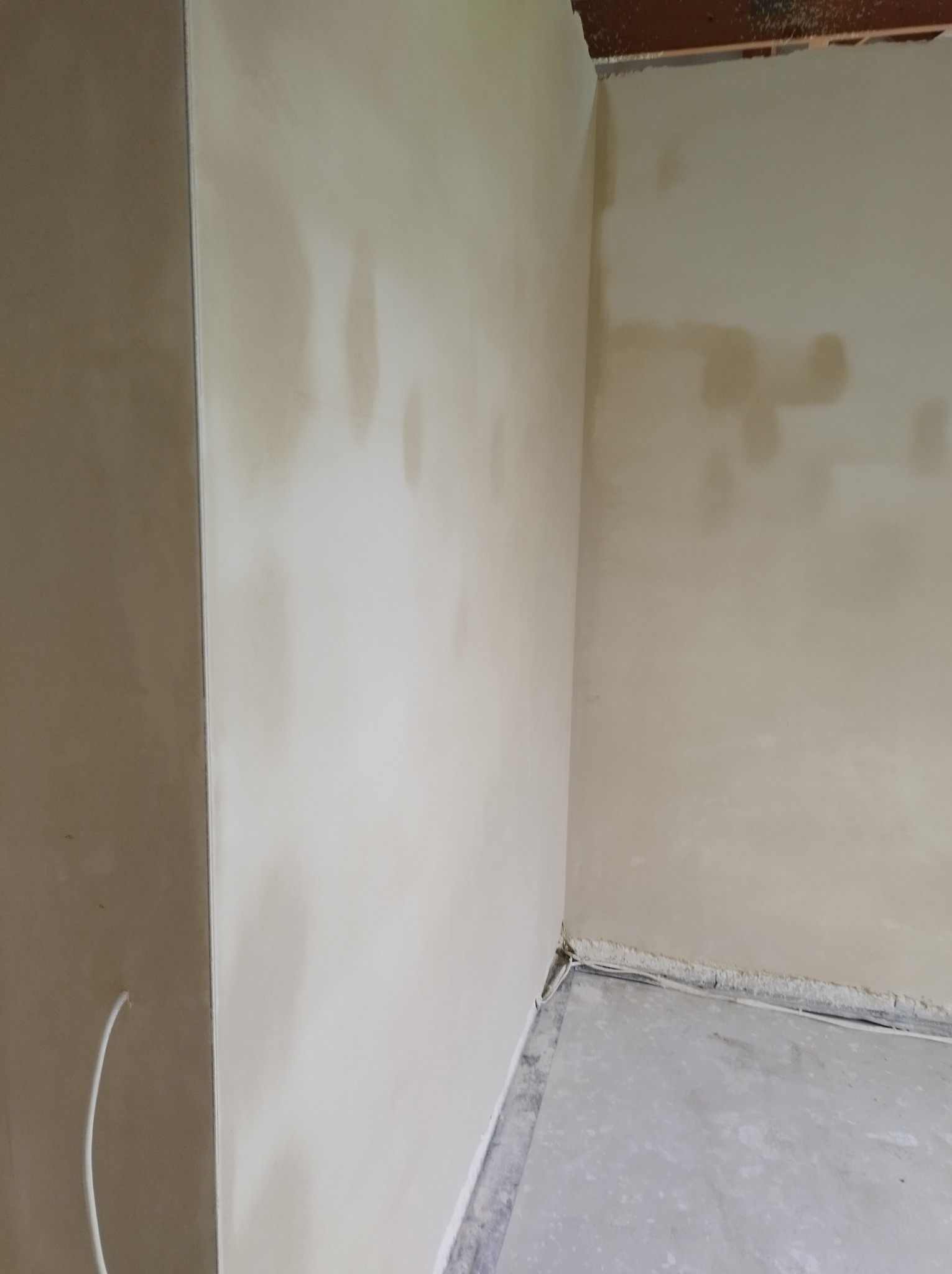 Świeżo nałożona, wilgotna, jasna warstwa na ścianach w rogu pomieszczenia, widoczne niedoskonałości i różnice w odcieniach, szare wykończenie podłogi.