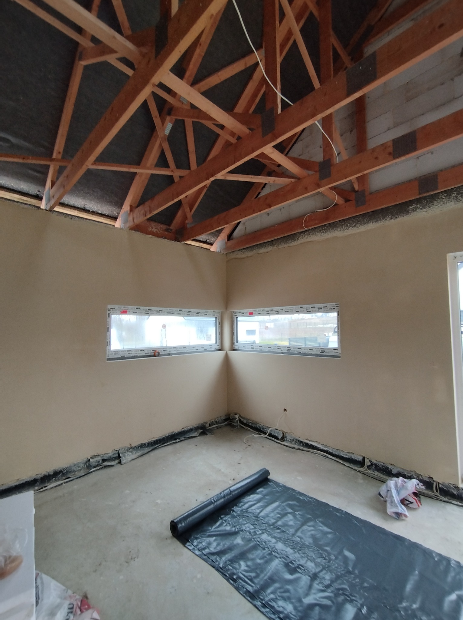 Surowe wnętrze z drewnianą konstrukcją dachu, świeżo wykończone ściany z dwoma poziomymi oknami, podłoga zabezpieczona czarną folią, widoczne ślady prac budowlanych.
