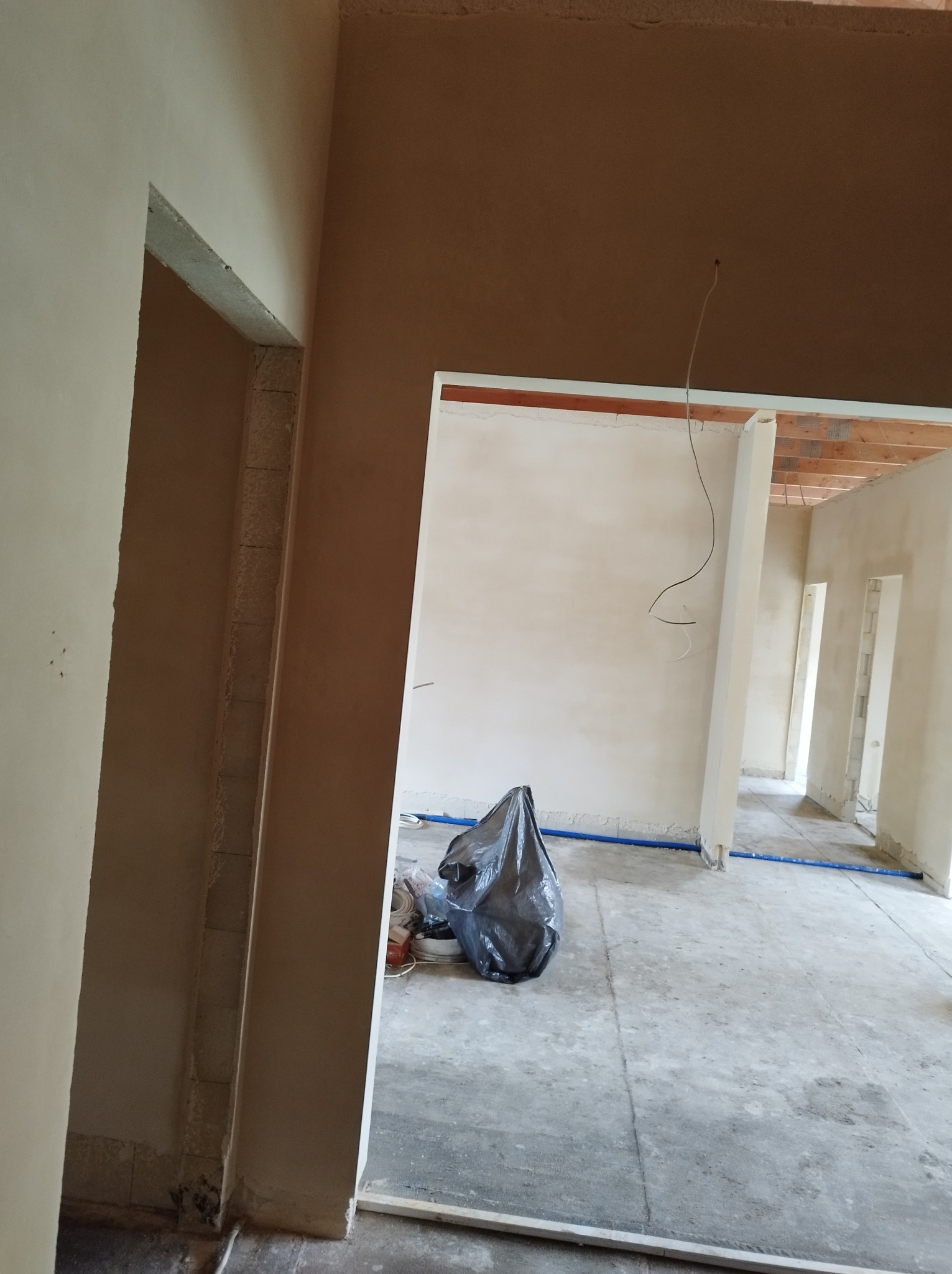 Wnętrze w trakcie prac wykończeniowych, widoczne ściany po nałożeniu tynku, podłoga betonowa z niebieskimi rurami, śmieci w czarnym worku, surowy stan z widoczną konstrukcją drewnianą stropu.
