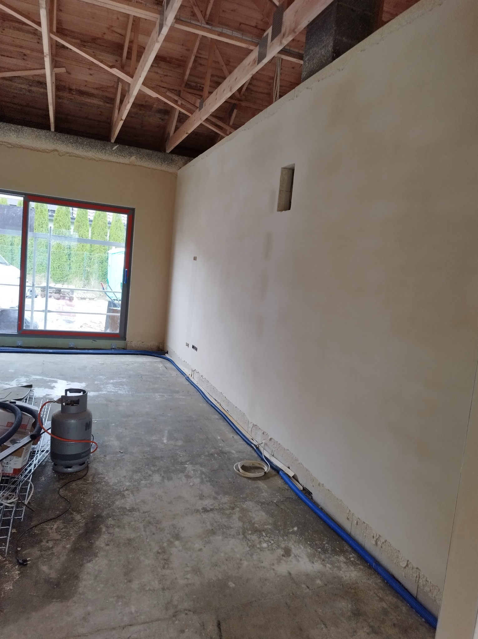 Wnętrze w trakcie prac wykończeniowych: ściana po nałożeniu tynku, widoczna instalacja elektryczna w peszlu przy podłodze, butla gazowa i surowy beton na podłodze.